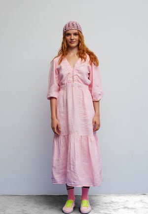 Woman with red hair wearing a pink midi dress, matching crocheted beanie, pink socks, and pink sneakers with neon green laces, standing against a plain wall.
