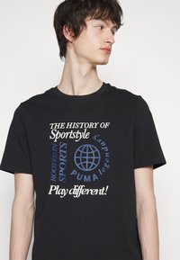 Zwarte katoenen t-shirt met witte en blauwe tekstgraphics, inclusief een wereldbol en zinnen zoals "DE GESCHIEDENIS VAN Sportsstyle" en "Speel anders!"