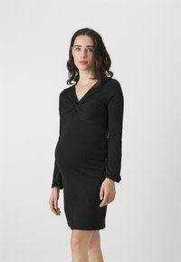 Juoda nėštumo suknelė, pritaikyta figūrai, ilgomis rankovėmis ir vingiuota priekyje, pagaminta iš tempimo audinio, iki vidurinės šlaunies.