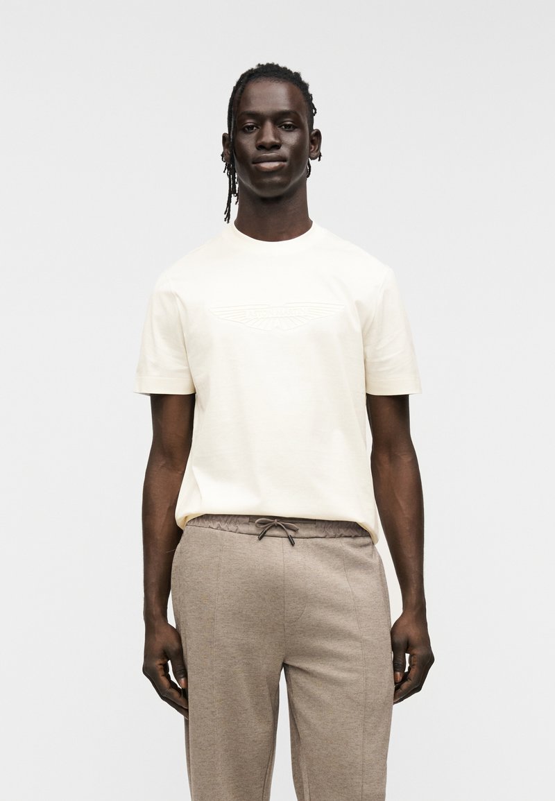 Κρεμ κοντομάνικο t-shirt με ανάγλυφο λογότυπο, συνδυασμένο με μπεζ παντελόνι με κορδόνι και μαλακό, ελαφρώς ραβδωτό ύφασμα.