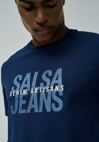 Camiseta azul marino hecha de algodón, con un atrevido texto gráfico "SALSA JEANS" y "DENIM ARTISANS" en azul claro y blanco.