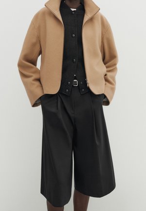 Person wearing beige zip-up jacket, black buttoned shirt, and loose black knee-length shorts with hands in pockets.