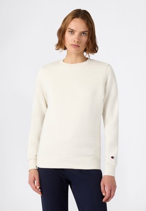 Junge Person mit kurzem Haar, die einen schlichten, ecrufarbenen Langarmpullover und dunkle Hosen vor einem weißen Hintergrund trägt.
