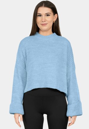 Frau trägt einen hellblauen grob gestrickten Pullover mit umgekrempelten Ärmeln und schwarzen Leggings, steht vor einem einfarbigen Hintergrund.