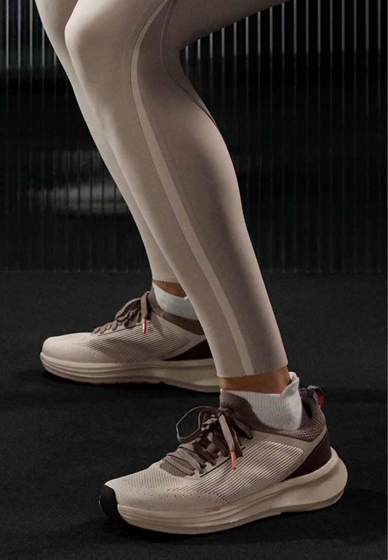 Jambes en leggings beiges et chaussettes blanches aux chevilles portant des chaussures de course beiges et marron à lacets sur un sol sombre.