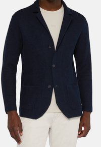 Námořnické modré pletené sako s klopovým límcem, dvěma předními kapsami a zapínáním na knoflík. Sako je kombinováno se svetlem barevným tričkem s kulatým výstřihem pod ním.