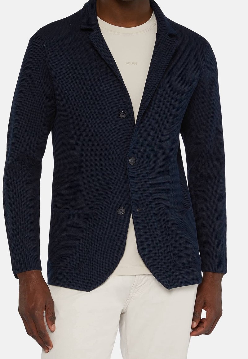 Námořnické modré pletené sako s klopovým límcem, dvěma předními kapsami a zapínáním na knoflík. Sako je kombinováno se svetlem barevným tričkem s kulatým výstřihem pod ním.