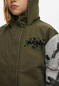 Olivgrön jacka med paljettbroderi av "Mickey", med en grå ärm med grafik av Mickey Mouse. Texturerat tyg, huva.