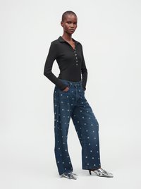 Femme portant un haut noir à manches longues, un jean large bleu avec des motifs œillets, et des talons hauts argentés pointus, debout, les mains dans les poches.