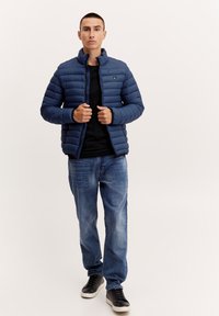 Marineblå polstret jakke med høy krage, med glidelås foran og ribbestrikkede ermer, kombinert med en svart topp og blå jeans. Svart sneakers fullfører antrekket.