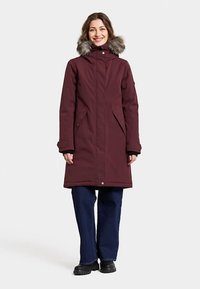 Maroon parka with a fur-lined hood, zippered front, side pockets, and a straight cut. Worn over dark blue pants and black boots.