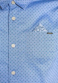 Camisa de color azul claro con pequeños lunares oscuros, diseño de botón, bolsillo en el pecho y un pañuelo de bolsillo a rayas en contraste, junto con el logo.