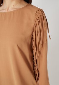 Primer plano de una blusa marrón de manga larga con detalles de flecos a lo largo de la manga exterior y un simple escote redondo.