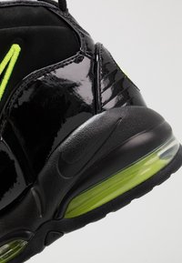 Černá sportovní bota s lesklou lakovou kůží, limetkově zelenými akcenty, texturovanou podrážkou a viditelným vzduchovým polštářem v patě.