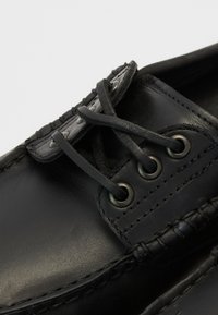 Chaussure en cuir noir avec un bout rond, des coutures visibles, un design à lacets et des œillets en métal. Texture lisse et éclat subtil.