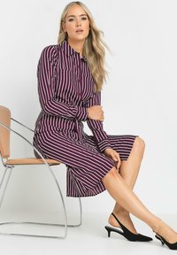 Abito a maniche lunghe color bordeaux con strisce verticali bianche, chiusura frontale a bottoni e gonna midi. Indossato con décolleté nere con cinturino alla caviglia.