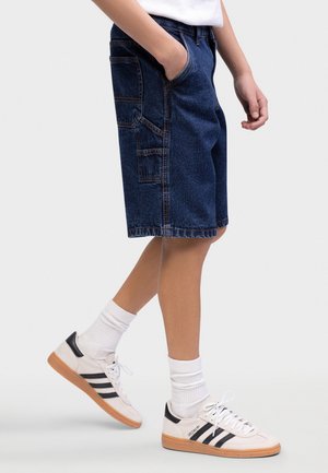 Personne portant un short en jean bleu foncé avec plusieurs poches, des chaussettes blanches et des baskets blanches avec des bandes noires, marchant sur un fond clair.