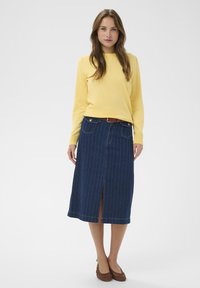 Pull jaune à manches longues associé à une jupe en denim bleu foncé avec des rayures verticales, une fente à l'avant, et des chaussures tressées marron.