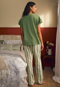 Sieviete stāv basām kājām guļamistabā, valkājot zaļu īsroku topu un baltas šauras ziedu raksta pidžamas bikses, blakus gultai un naktsgaldiņam ar ziediem.