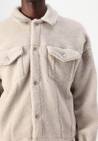 Beige fleece jack met een textuur van pluche. Voorzien van drukknoopsluiting en twee voorkruiszakken, wat bijdraagt aan een ontspannen pasvorm.