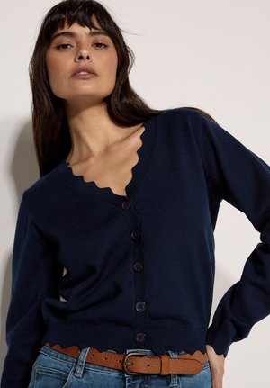 Donna che indossa un cardigan blu navy con bordi smerlati e bottoni scuri, infilato in jeans blu con cintura marrone, che posa con una mano sul fianco.