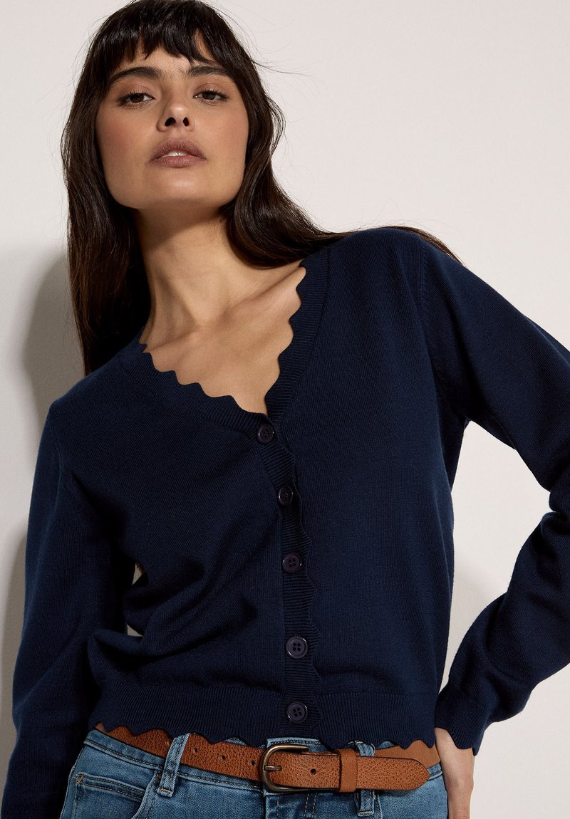 Femme portant un cardigan bleu marine à bord festonné avec des boutons foncés, rentré dans un jean bleu avec une ceinture marron, posant avec une main sur la hanche.