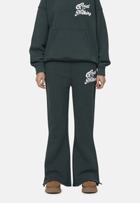 Ciemnozielony sweatshirt i pasujące spodnie z miękkiego materiału. Sweatshirt posiada kieszeń z przodu i biały nadruk; spodnie mają rozszerzane nogawki i nadruki po bokach.