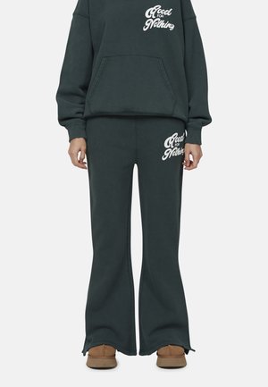 Felpa verde scuro e pantaloni coordinati in tessuto morbido. La felpa presenta una tasca frontale e testo bianco; i pantaloni hanno gambe svasate e stampe sui lati.