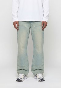 Άνδρας που φοράει φαρδιά τζιν σε ανοιχτό γαλάζιο, λευκό πουκάμισο με μακριά μανίκια και λευκά αθλητικά παπούτσια, στέκεται μπροστά σε μια απλή φόντο.