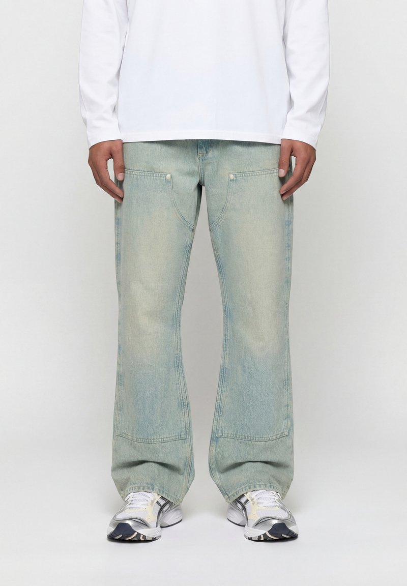 Άνδρας που φοράει φαρδιά τζιν σε ανοιχτό γαλάζιο, λευκό πουκάμισο με μακριά μανίκια και λευκά αθλητικά παπούτσια, στέκεται μπροστά σε μια απλή φόντο.