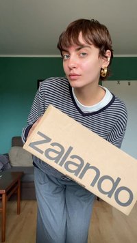 Brown shipping box with "zalando" in bold gray text, held by a person in a striped navy and white sweater and gray trousers.