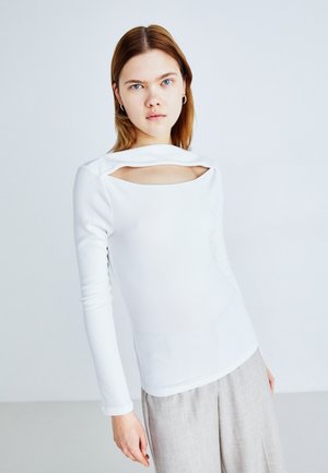 White ribbed long-sleeve top with a cut-out detail at the neckline and a fitted silhouette, paired with light gray pants.