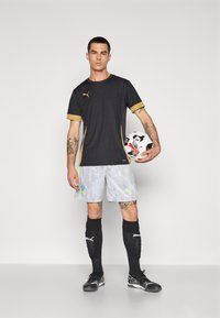 Sort atletisk skjorte med guld accenter og grå mønstrede shorts. Modellen holder en fodbold og har sorte sokker og støvler på.