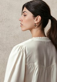 Lichtromige blouse met opbollende mouwen, gerimpelde halslijn en een soepele textuur. Heeft een subtiele gathering bij de achternaad.