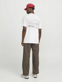 Hvid t-shirt med et grafik på ryggen, der står "Track Club." Båret med brune brede bukser og en rød kasket. Sportssko synlige.