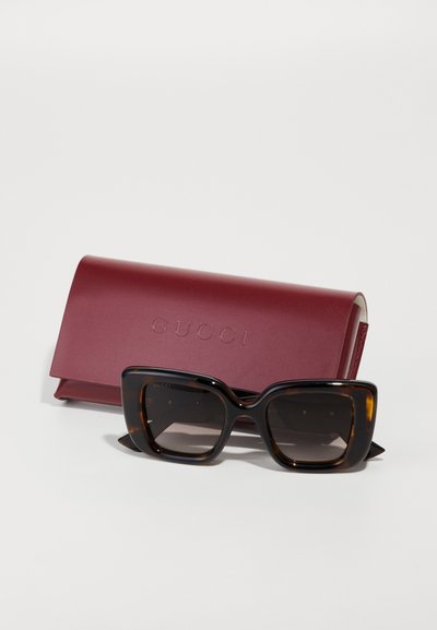 Gucci zonnebril met oversized donkere schildpadvormige monturen en vervagende lenzen, vergezeld van een bordeauxleren hoes met de opdruk "GUCCI."