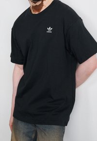 Osoba oblečená v černém tričku Adidas s bílým logem na hrudi a vybledlých modrých džínech, stojí proti bílé zdi.