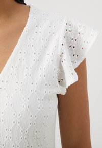 Vit texturerad topp med v-ringning och korta, volangärmar. Har intrikata broderade mönster och en något genomskinlig framtoning.