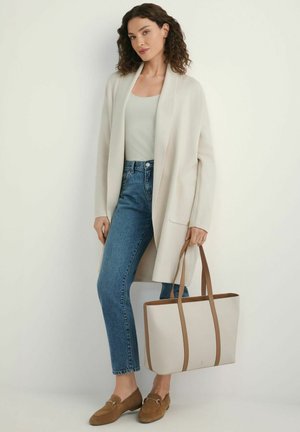 Kobieta ubrana w kremowy kardigan, jasną bluzkę, niebieskie dżinsy, brązowe mokasyny, trzymająca beżową torbę na ramię z brązowymi rączkami, stojąca na tle białej ściany.