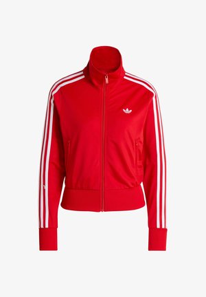 Röd jacka med dragkedja och hög krage, vitt mönster med tre ränder på ärmarna och liten vit logga på bröstet. Slät tygstruktur.