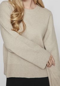 Beige langärmliger Strickpullover mit Rundhalsausschnitt, gerippten Bündchen und einem leicht verlängerten hinteren Saum. Weicher Stoff mit minimalem Muster.