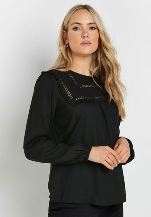 Blusa negra de manga larga con un cuello con volantes y costuras decorativas. Tejido suave con un corte relajado y puños fruncidos.