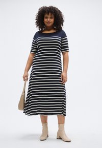 Tengerészkék és krémszínű csíkos midi ruha rövid ujjakkal, csónaknyakkal és laza szabással. Bézs bokacsizmával viselve, kézben egy barna táskával.