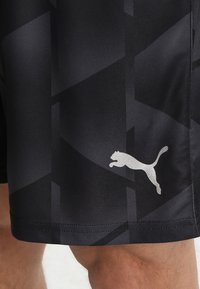 Svarta sportshorts i slät tyg, med ett geometriskt mönster i mörka nyanser och en silverfärgad Puma-logotyp på undersidan.