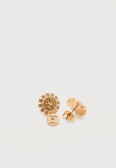 Brincos redondos em tom dourado com pedras claras em redor de um emblema de logótipo em relevo, exibidos sobre um fundo branco.