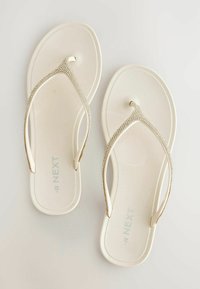 Tongs plates blanches avec des brides argentées scintillantes et le logo "NEXT" sur la semelle, présentées sur un fond clair uni.