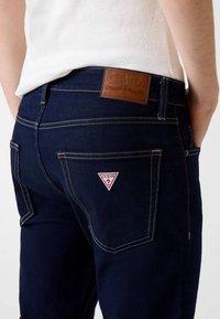 Jeans in denim blu scuro con cuciture gialle. Presentano una patch in pelle marrone e un logo Guess rosso e bianco sulla tasca posteriore.