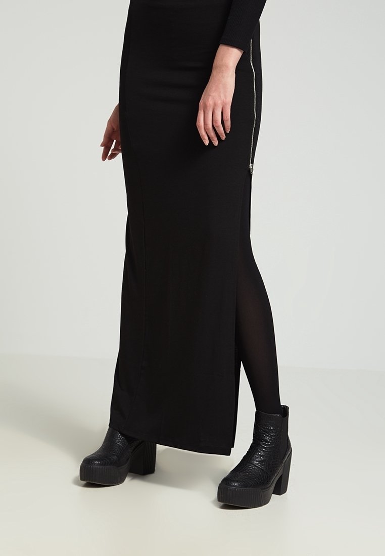 Jupe maxi noire avec une fermeture éclair latérale, une texture lisse et une silhouette droite. Associée à des bottes plateformes noires texturées.