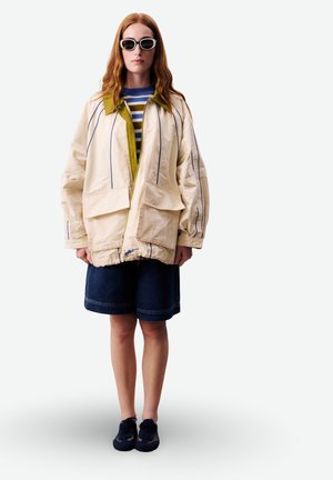 Vrouw met lang rood haar die een witte oversized jas met blauwe strepen draagt, een gestreept shirt, donkere shorts, zwarte sneakers en een witte zonnebril.