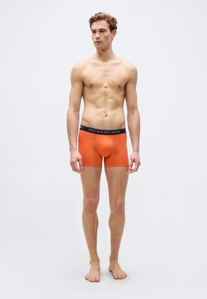 Junger Mann steht barfuß da, trägt orangefarbene Polo Ralph Lauren Boxershorts mit einem schwarzen Bund, vor einem schlichten hellen Hintergrund.